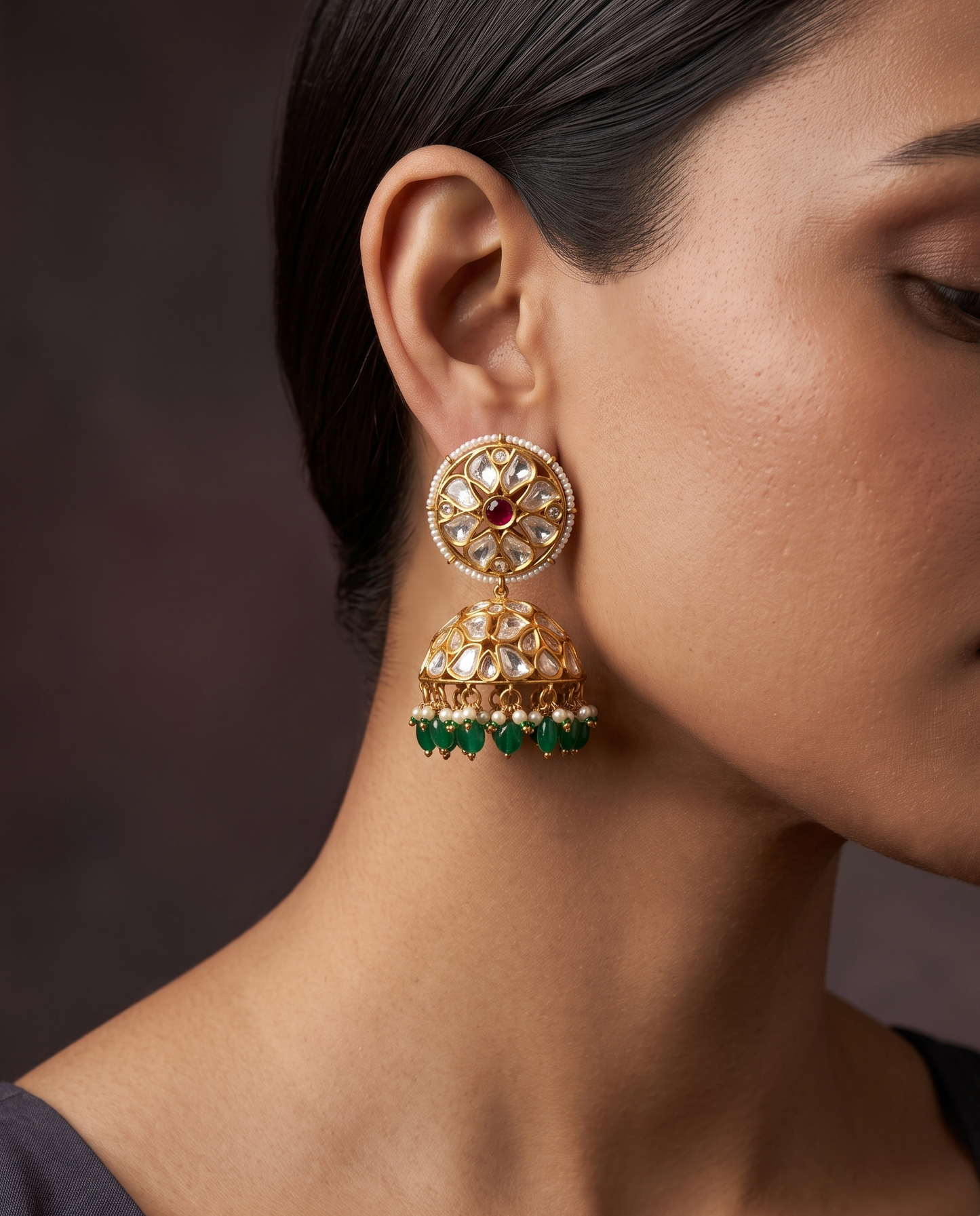 A close-up of a woman wearing a large gold-toned traditional Indian jhumka earring with a red stone, white stones, and green bead drops, set against a dark brown background.