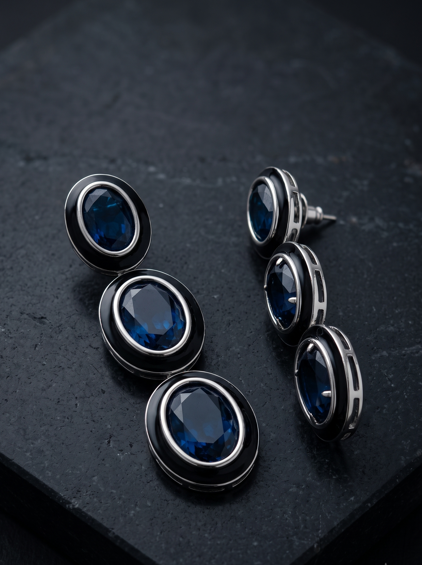 A pair of oval drop earrings with deep blue faceted gemstones surrounded by black and silver-toned metal settings, displayed on a dark textured stone surface.