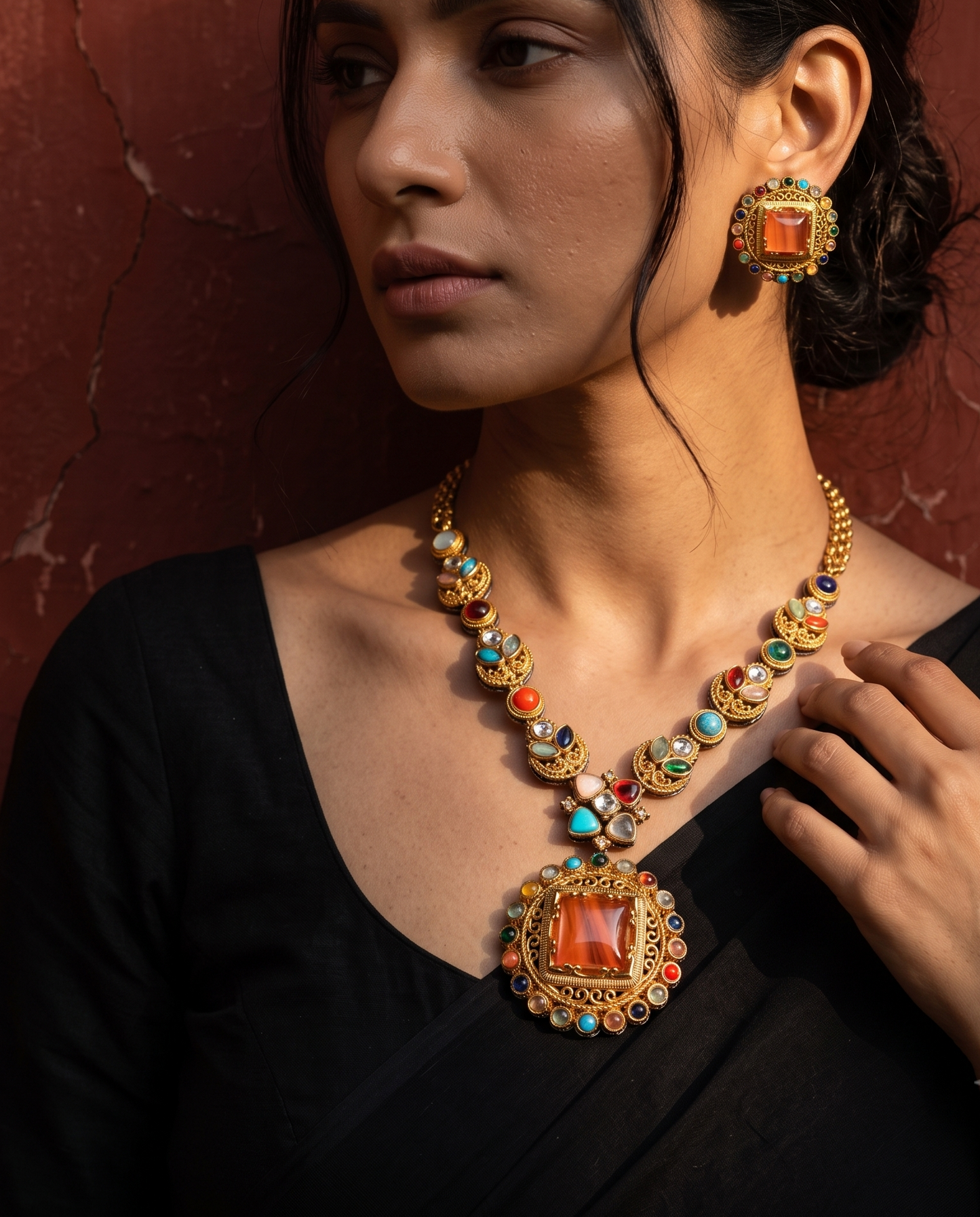 A woman in a black blouse models an ornate gold-toned jewelry set featuring a necklace and matching earring, both decorated with colorful stones and a large orange-red central gem, set against a textured reddish-brown wall.