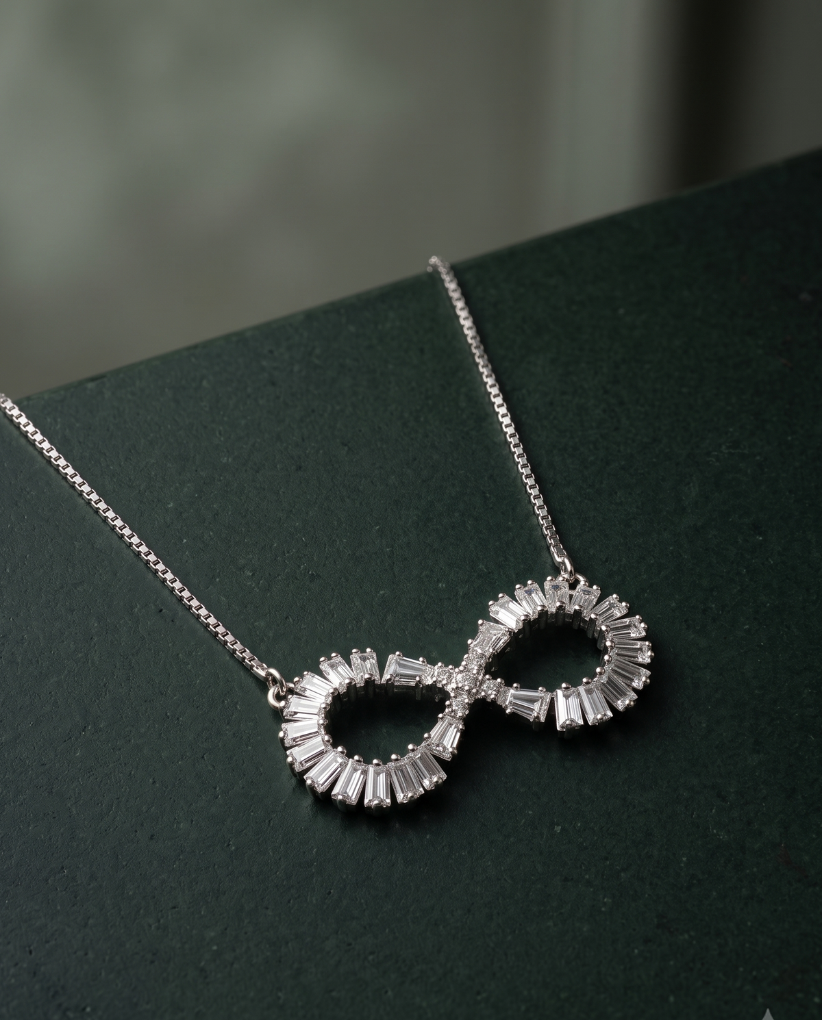 A silver infinity-shaped pendant necklace adorned with baguette-cut clear stones, resting on a dark green textured surface.