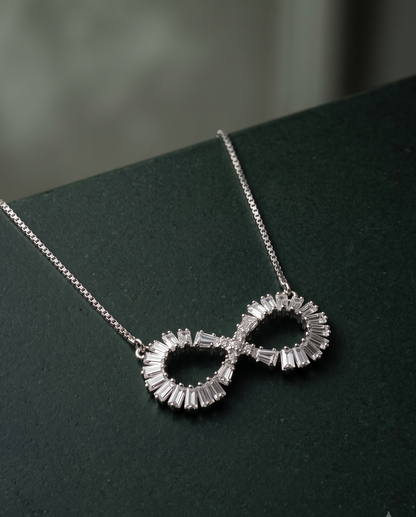 A silver infinity-shaped pendant necklace adorned with baguette-cut clear stones, resting on a dark green textured surface.