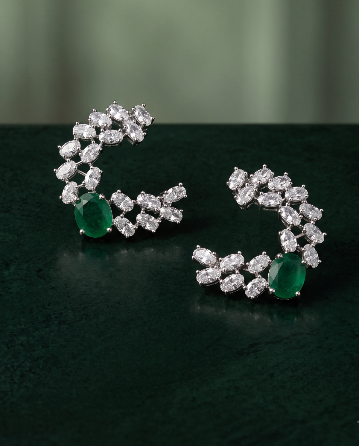 A pair of crescent-shaped silver-toned earrings set with clear stones and green emerald-cut accents, displayed on a dark green surface with a blurred green background.
