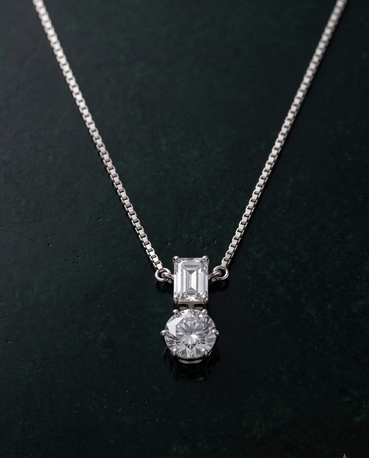 A silver-toned necklace with a two-stone pendant, featuring an emerald-cut clear gemstone above a round brilliant-cut clear gemstone, displayed on a dark textured background.