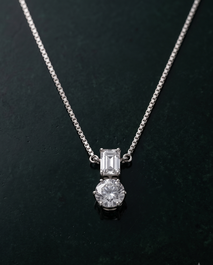 A silver-toned necklace with a two-stone pendant, featuring an emerald-cut clear gemstone above a round brilliant-cut clear gemstone, displayed on a dark textured background.