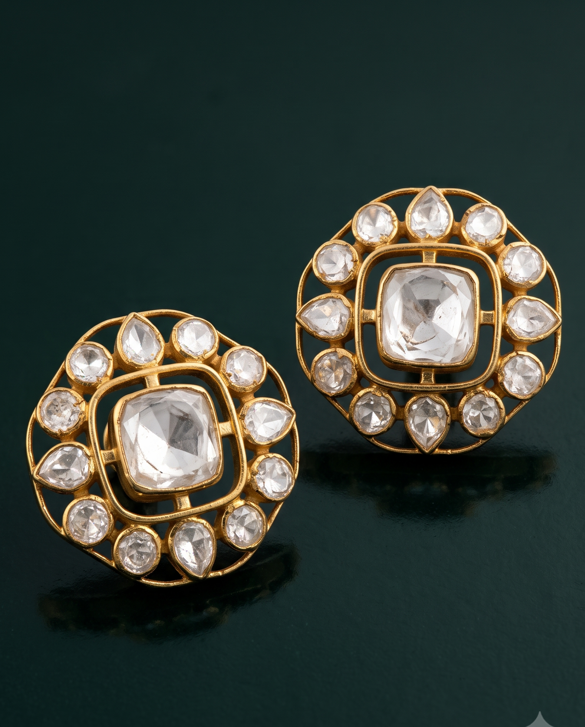 A pair of gold-toned square stud earrings with clear gemstones, displayed on a dark green reflective surface.