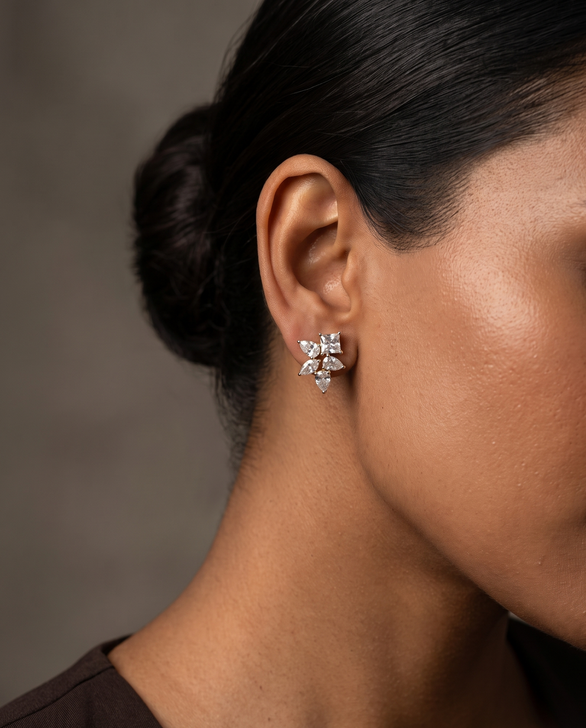 A close-up side profile of a woman wearing a small silver-toned flower-shaped stud earring with clear stones, against a soft warm gray background.