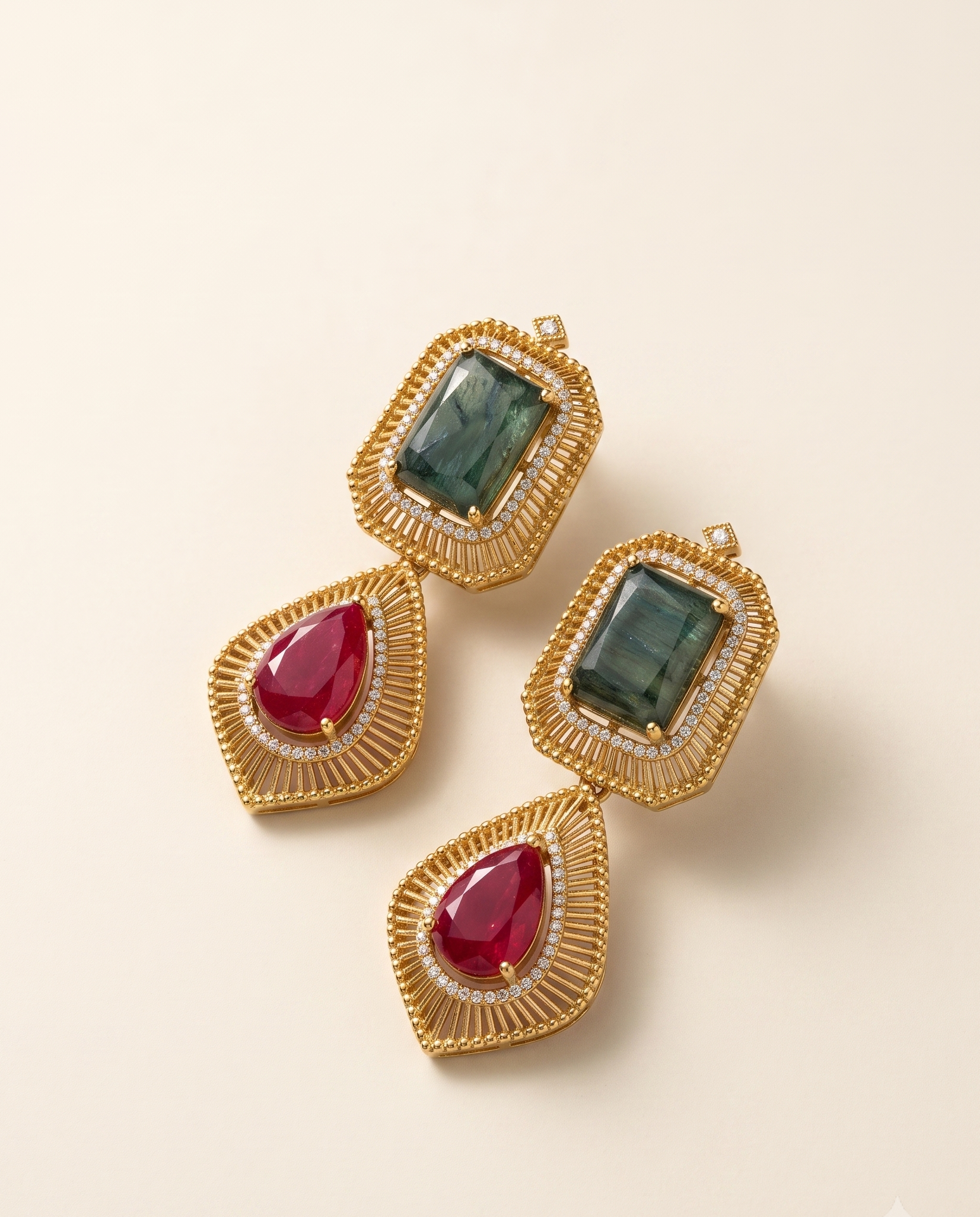 A pair of gold-toned statement earrings with rectangular green gemstones in the top sections and teardrop-shaped red gemstones in the lower sections, accented with small clear stones, photographed on a warm off-white background.