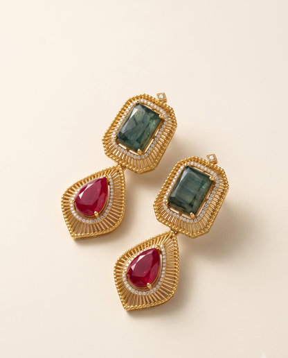 A pair of gold-toned statement earrings with rectangular green gemstones in the top sections and teardrop-shaped red gemstones in the lower sections, accented with small clear stones, photographed on a warm off-white background.