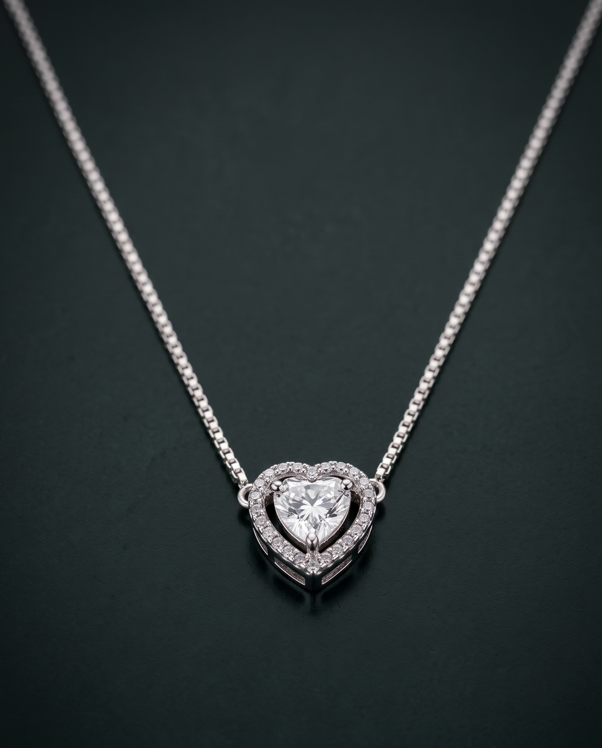 A silver heart pendant necklace with a double-heart halo design and a central heart-shaped clear gemstone, set against a dark green background.