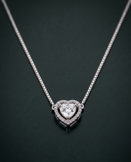 A silver heart pendant necklace with a double-heart halo design and a central heart-shaped clear gemstone, set against a dark green background.