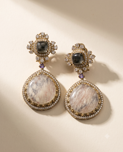 A pair of ornate gold-toned drop earrings with large oval pinkish-white marbled stone centers, surrounded by small clear stones and purple accent stones, photographed on a warm cream background.