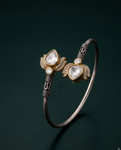 An ornate silver-toned bangle bracelet with two gold-toned decorative ends, each featuring a pear-shaped clear stone and small clear stones, photographed on a dark green background.
