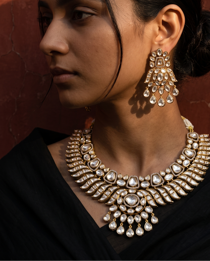 A woman in profile wearing an ornate gold-toned necklace and matching drop earrings with clear stones, set against a warm reddish-brown textured wall.