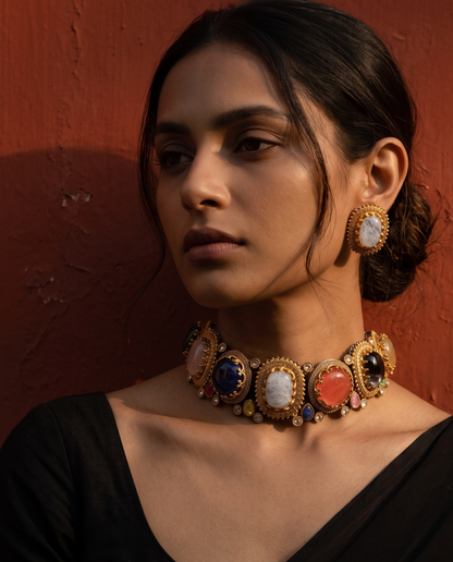 A woman wearing an ornate gold-toned choker necklace with large multicolored stone panels and matching drop earrings, posed against a textured reddish-brown wall.