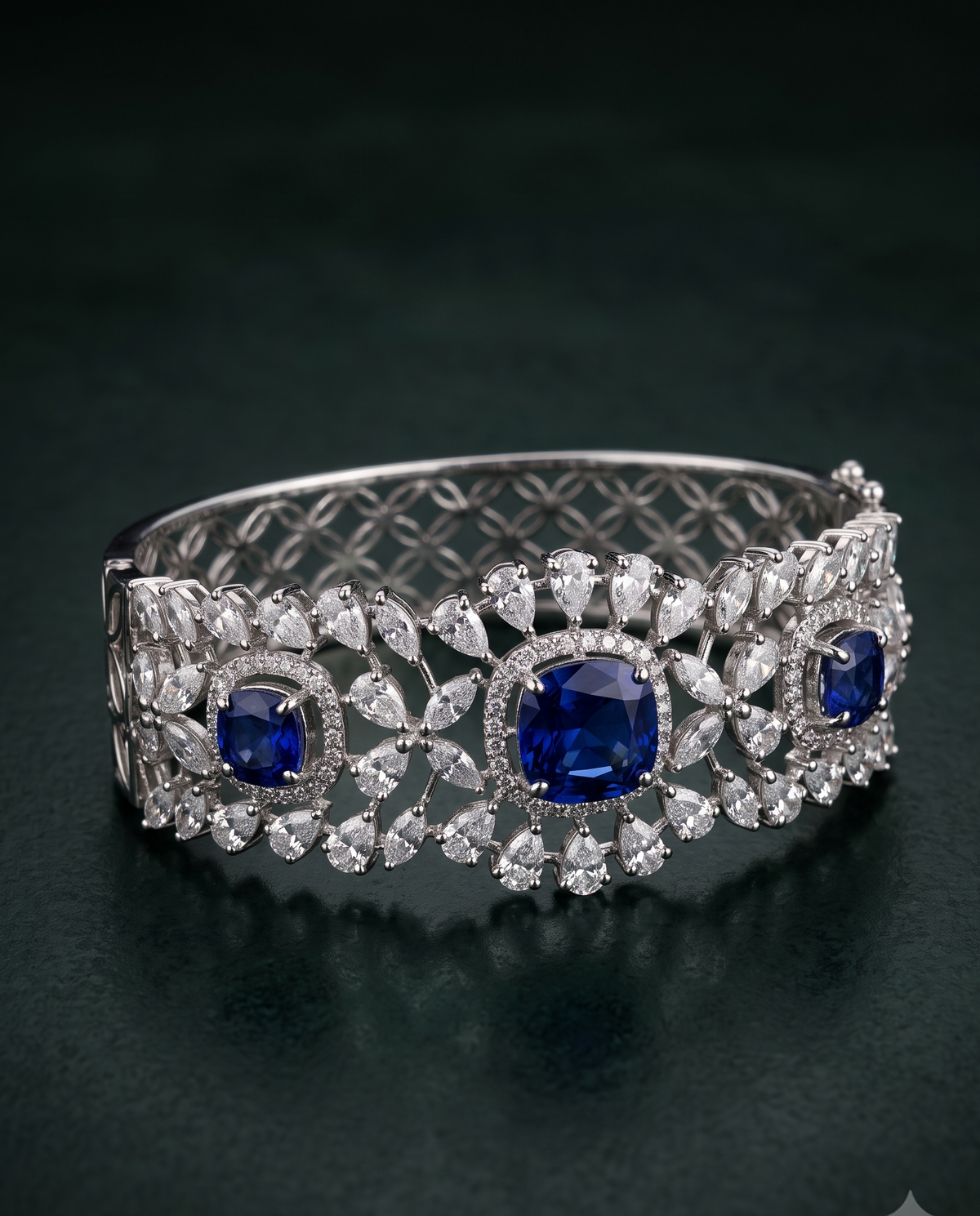 An ornate silver-toned cuff bracelet featuring three large round-cut deep blue sapphires and multiple surrounding clear stones, displayed on a dark green reflective surface.