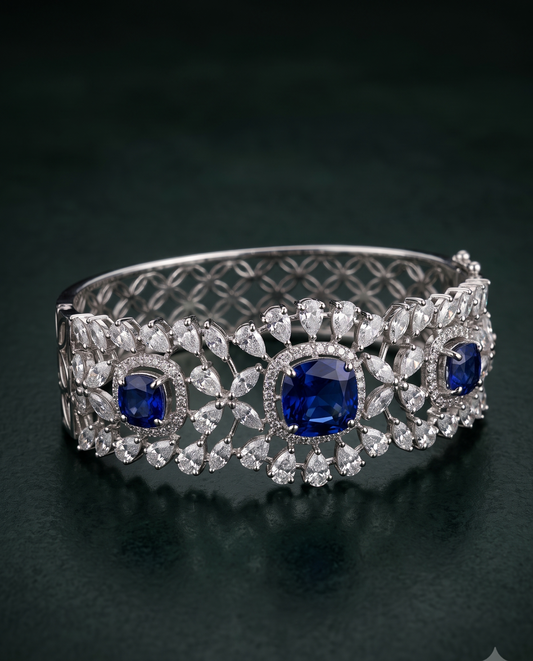 An ornate silver-toned cuff bracelet featuring three large round-cut deep blue sapphires and multiple surrounding clear stones, displayed on a dark green reflective surface.