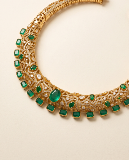 An ornate gold-toned choker necklace embellished with multiple emerald-cut green gemstones and clear stones, photographed on a clean, light beige background.