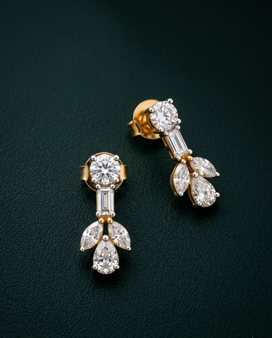 A pair of gold drop earrings with clear gemstones, including a round center stone, baguette side stones, and pear and marquise-shaped drops, displayed on a dark green textured background.