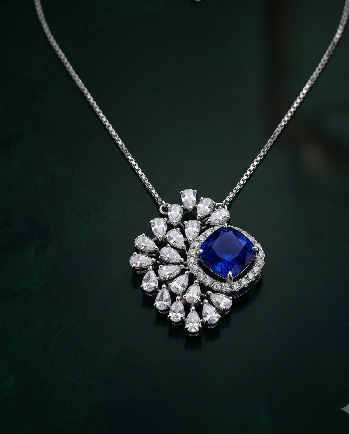 A silver-tone pendant necklace featuring a central deep blue cushion-cut gemstone surrounded by a halo of small clear stones, with a larger floral cluster of clear teardrop and round stones, displayed on a dark green background.