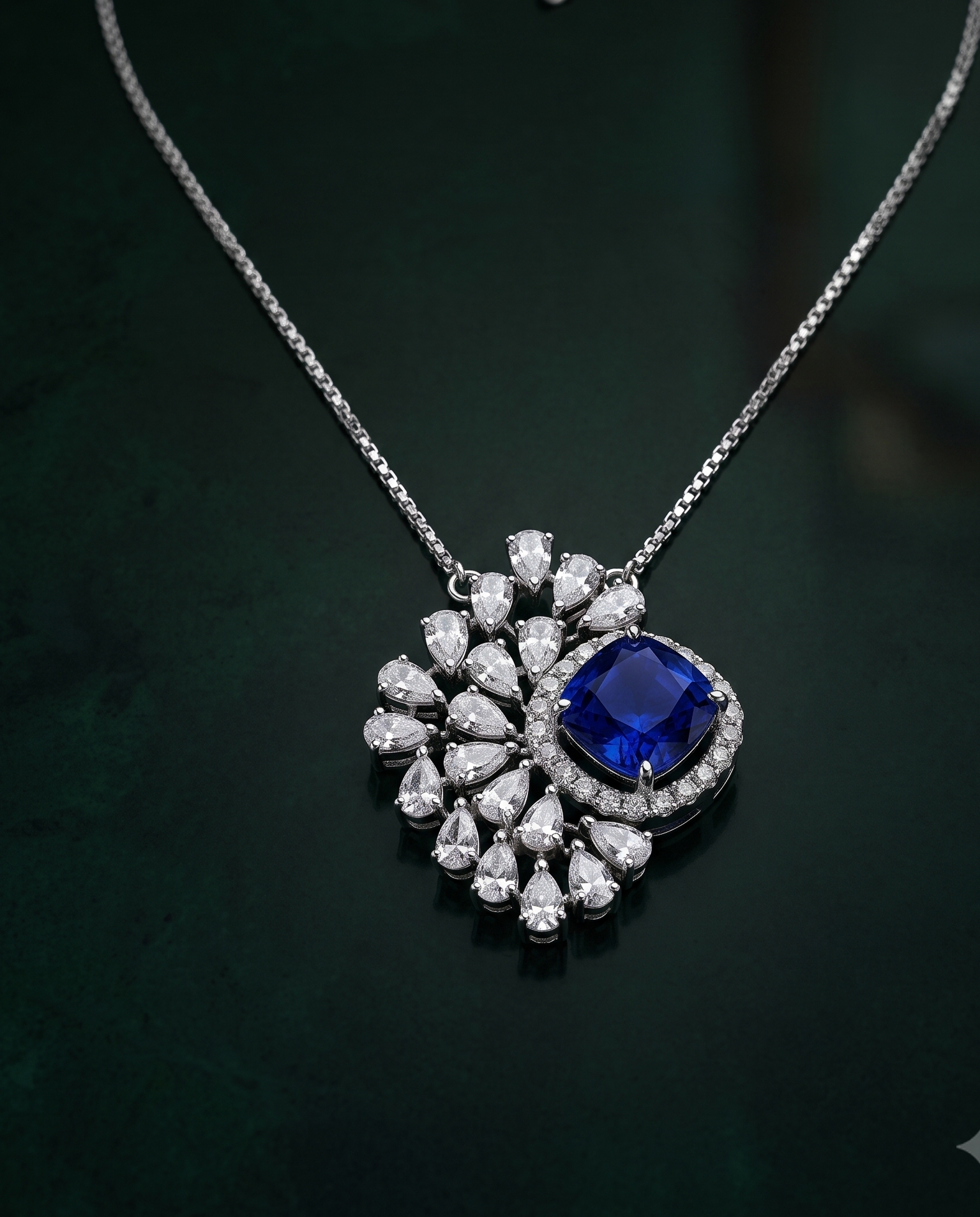 A silver-tone pendant necklace featuring a central deep blue cushion-cut gemstone surrounded by a halo of small clear stones, with a larger floral cluster of clear teardrop and round stones, displayed on a dark green background.