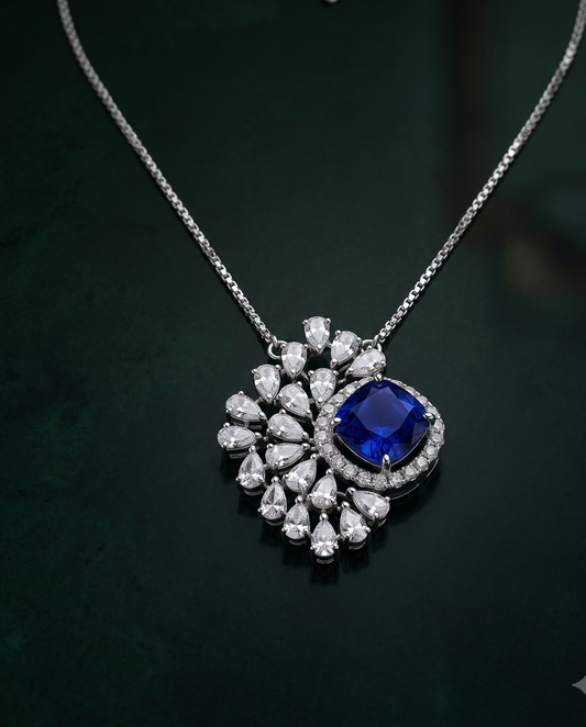 A silver-tone pendant necklace featuring a central deep blue cushion-cut gemstone surrounded by a halo of small clear stones, with a larger floral cluster of clear teardrop and round stones, displayed on a dark green background.