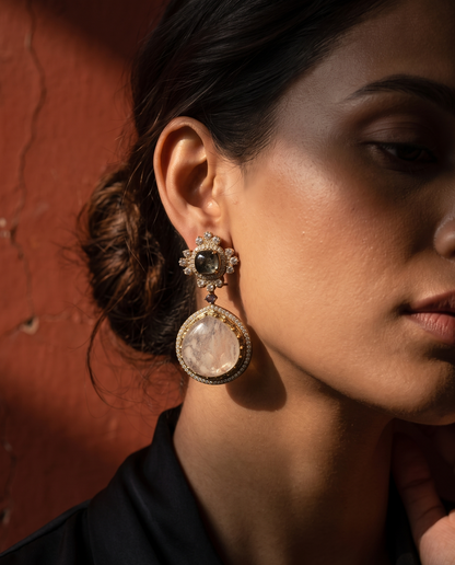 A close-up lifestyle shot of a woman wearing a large, ornate gold dangle earring with a milky white teardrop-shaped stone and a dark green round stone at the top, set against a textured terracotta wall.