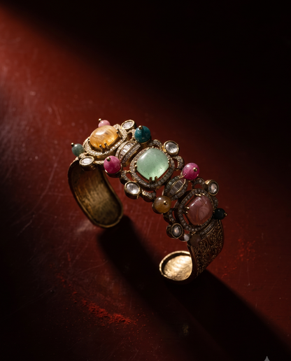 An ornate open cuff bracelet with a gold-toned base, decorated with multiple colorful gemstones including a light green center stone, pink, orange, and blue stones, photographed on a dark reddish-brown surface with dramatic lighting.
