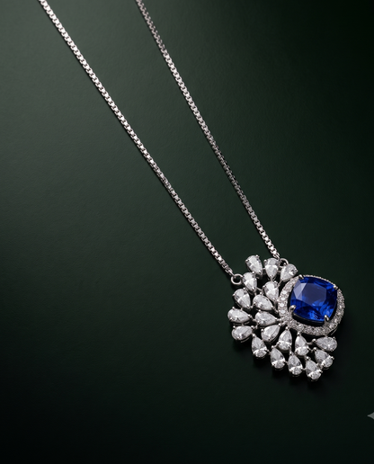 A luxury pendant necklace featuring a large oval-cut blue sapphire surrounded by a halo of small clear pavé stones on a silver-toned chain, set against a dark green gradient background.
