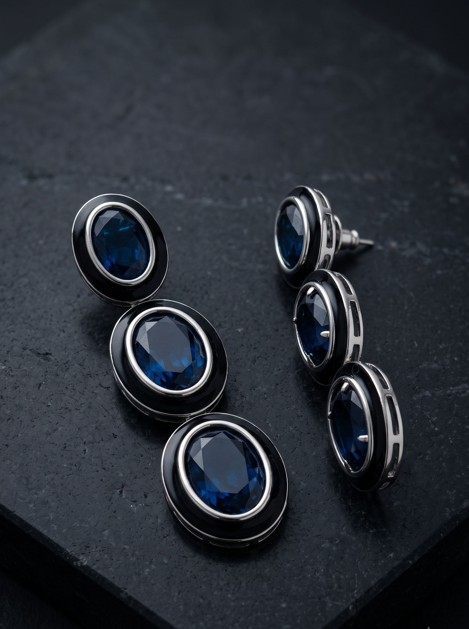 A pair of oval drop earrings with deep blue faceted gemstones surrounded by black and silver-toned metal settings, displayed on a dark textured stone surface.
