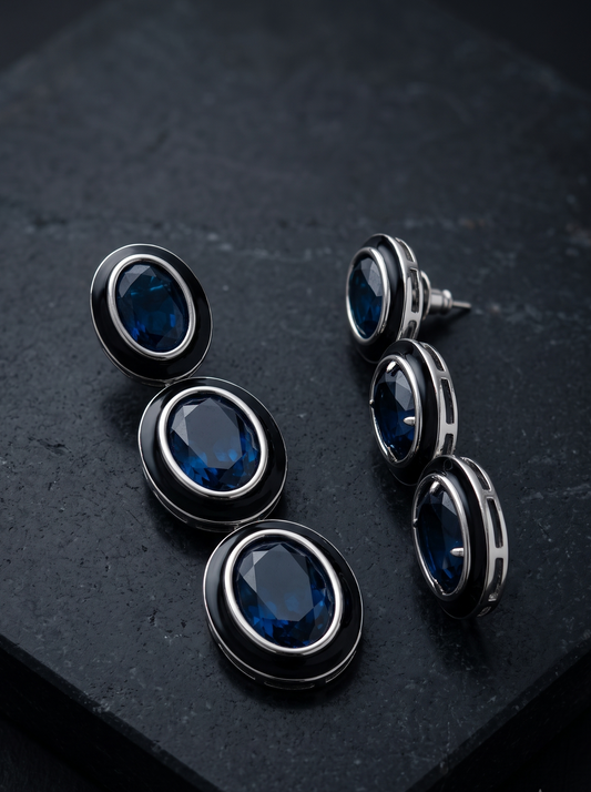 A pair of oval drop earrings with deep blue faceted gemstones surrounded by black and silver-toned metal settings, displayed on a dark textured stone surface.